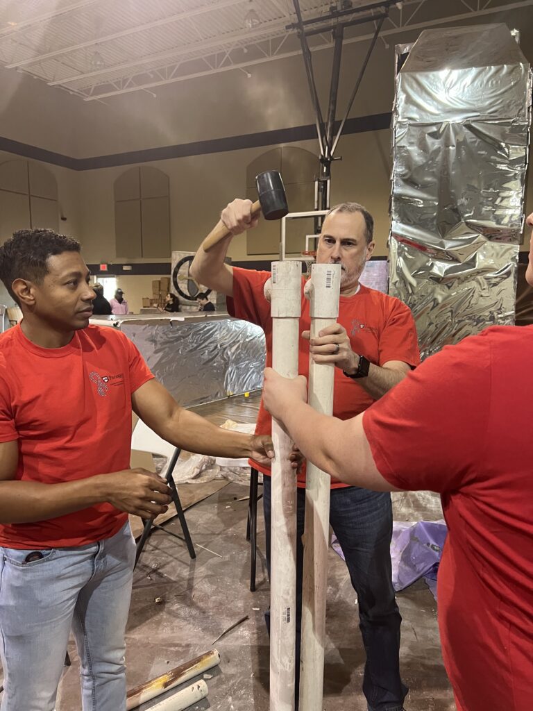 A group of bank employees in matching t-shirts are hammering lengths of pipe into each other. 